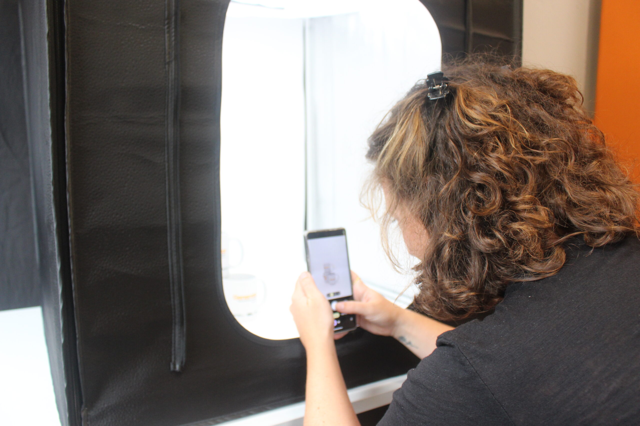 A woman using a smartphone to take a picture of some mugs that are inside a light-tent. The screen of the phone can be seen, and the back of the woman's head 