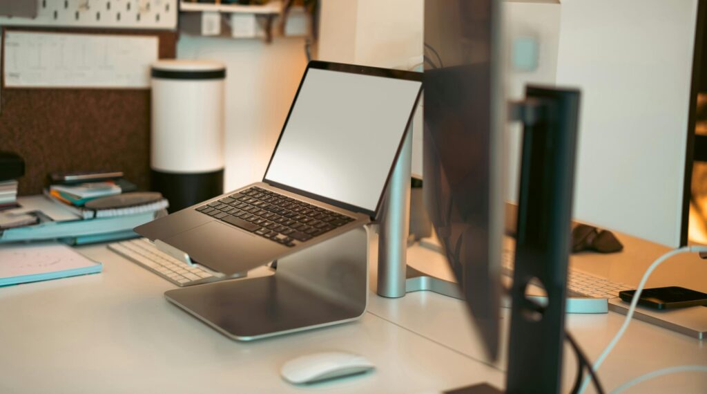 A desk with a laptop stand holding a laptop, a mouse and a desktop screen. The background is of a general home office.