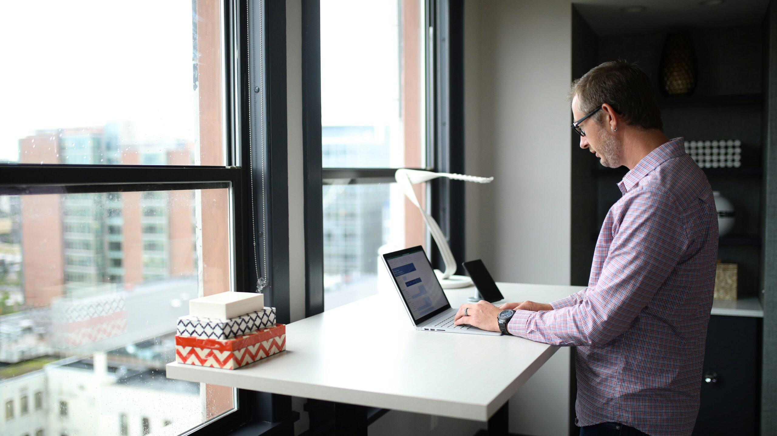 A white man standing at a desk in front of a large window overlooking city buildings